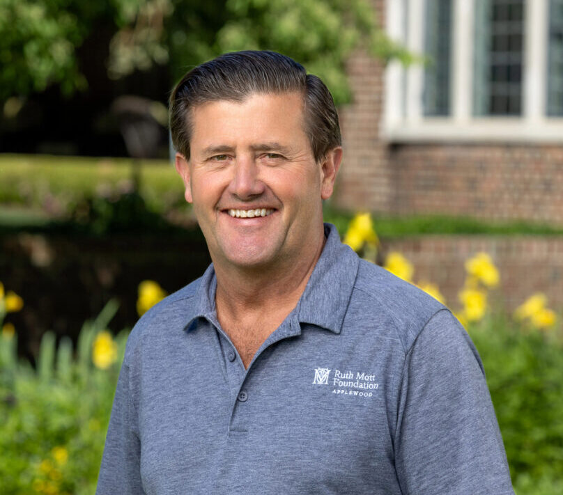 A man in a blue polo shirt stands outside in front of a brick building and green garden, smiling at the camera.