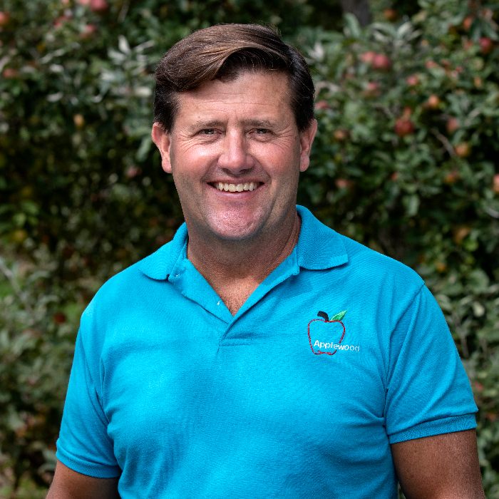 A man in a blue shirt standing in front of an apple tree.