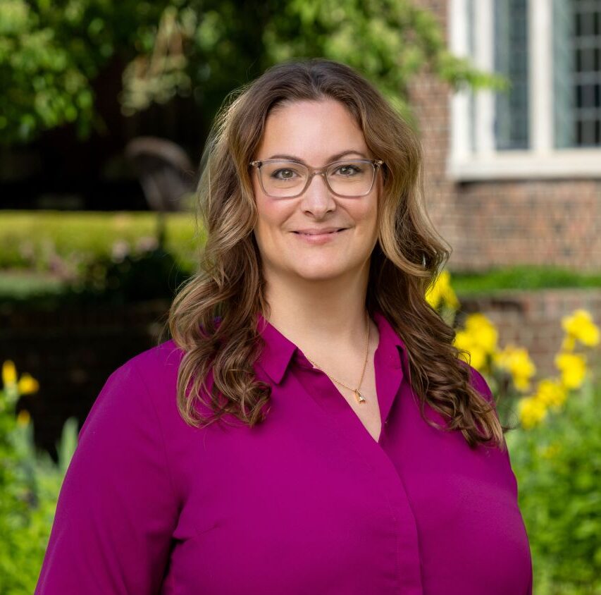 Woman with wavy brown hair and glasses wearing a magenta blouse stands outdoors in front of a garden and brick building.