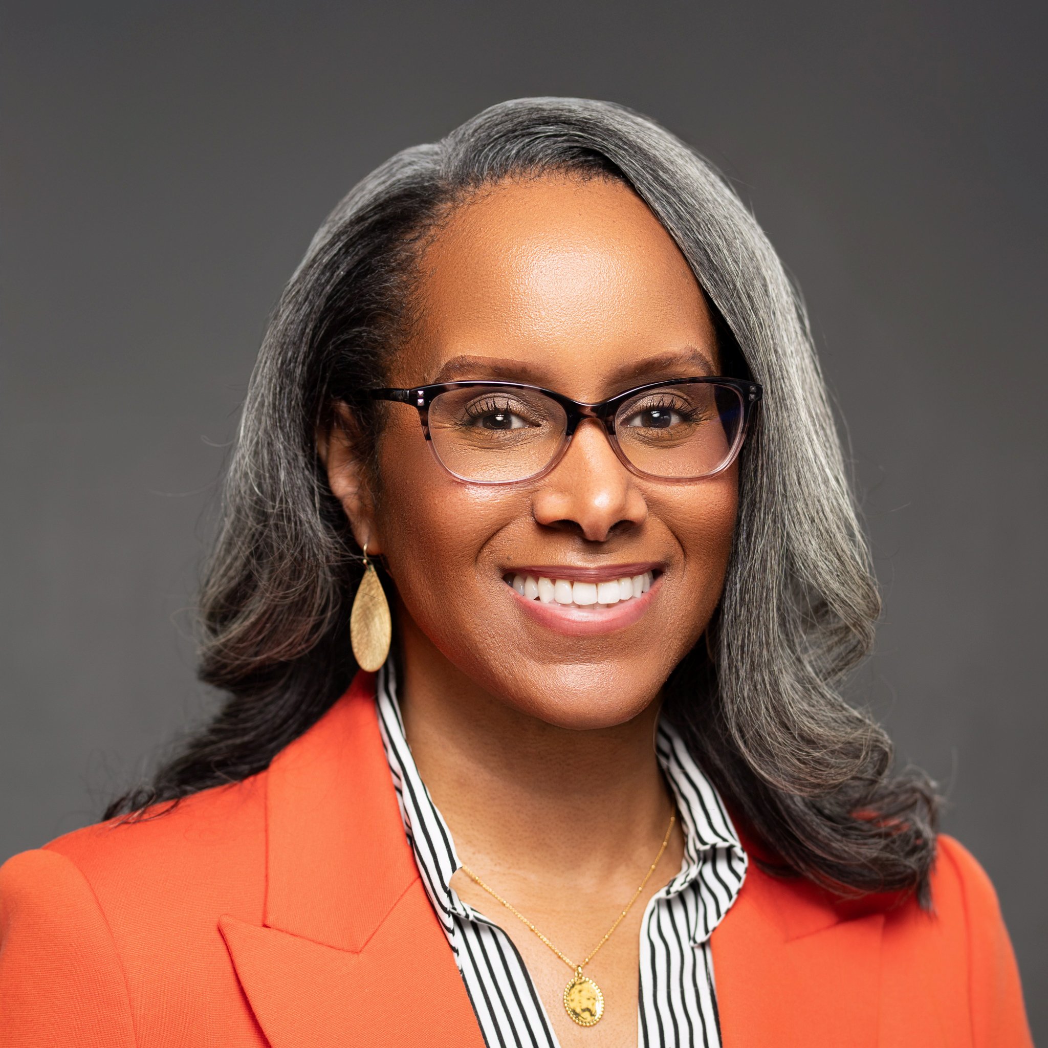 Smiling woman with glasses and long gray hair, wearing a coral blazer, striped blouse, gold necklace, and earrings, posed against a plain dark background.