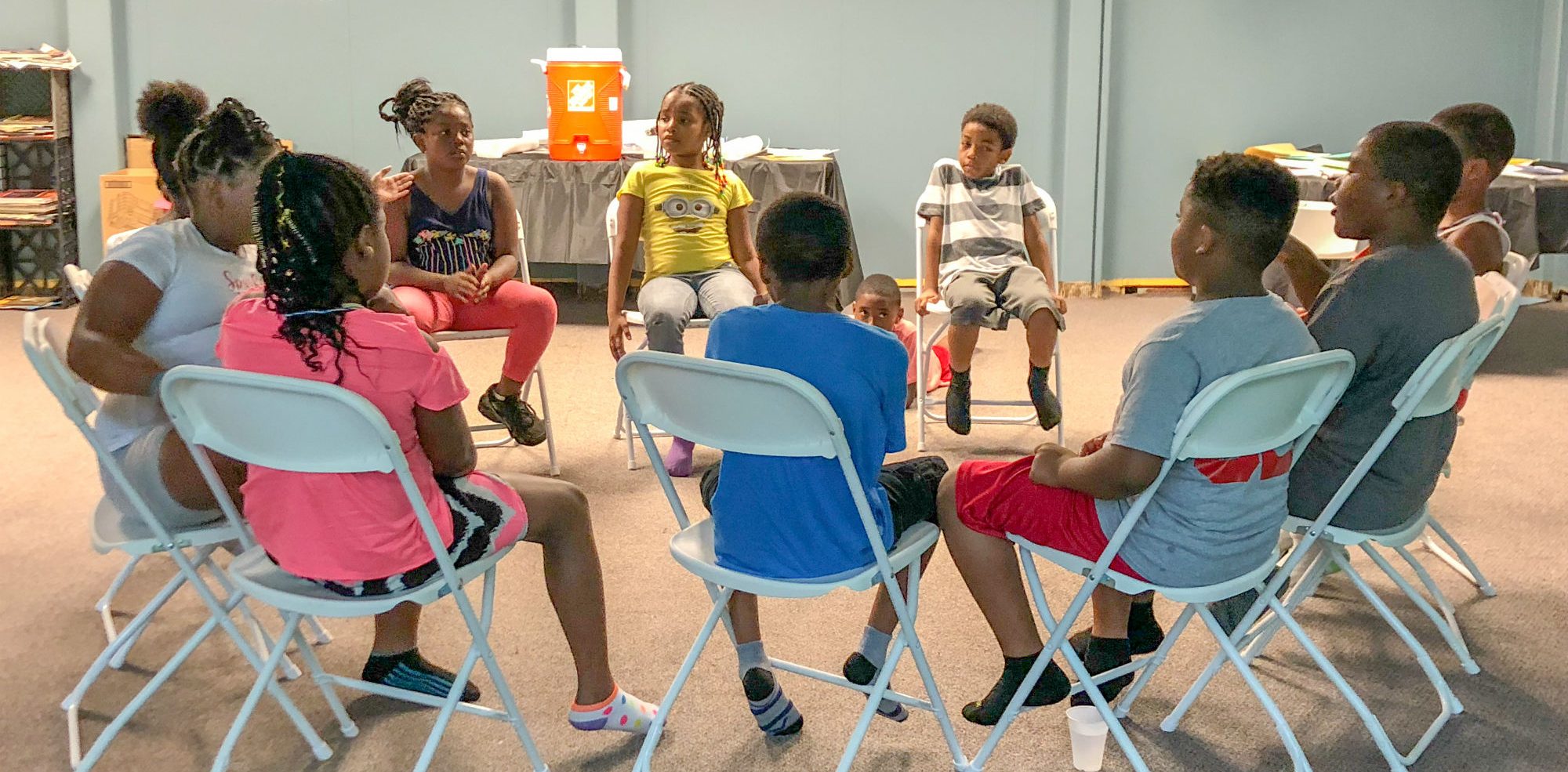 Children sitting in a circle on chairs inside a building.