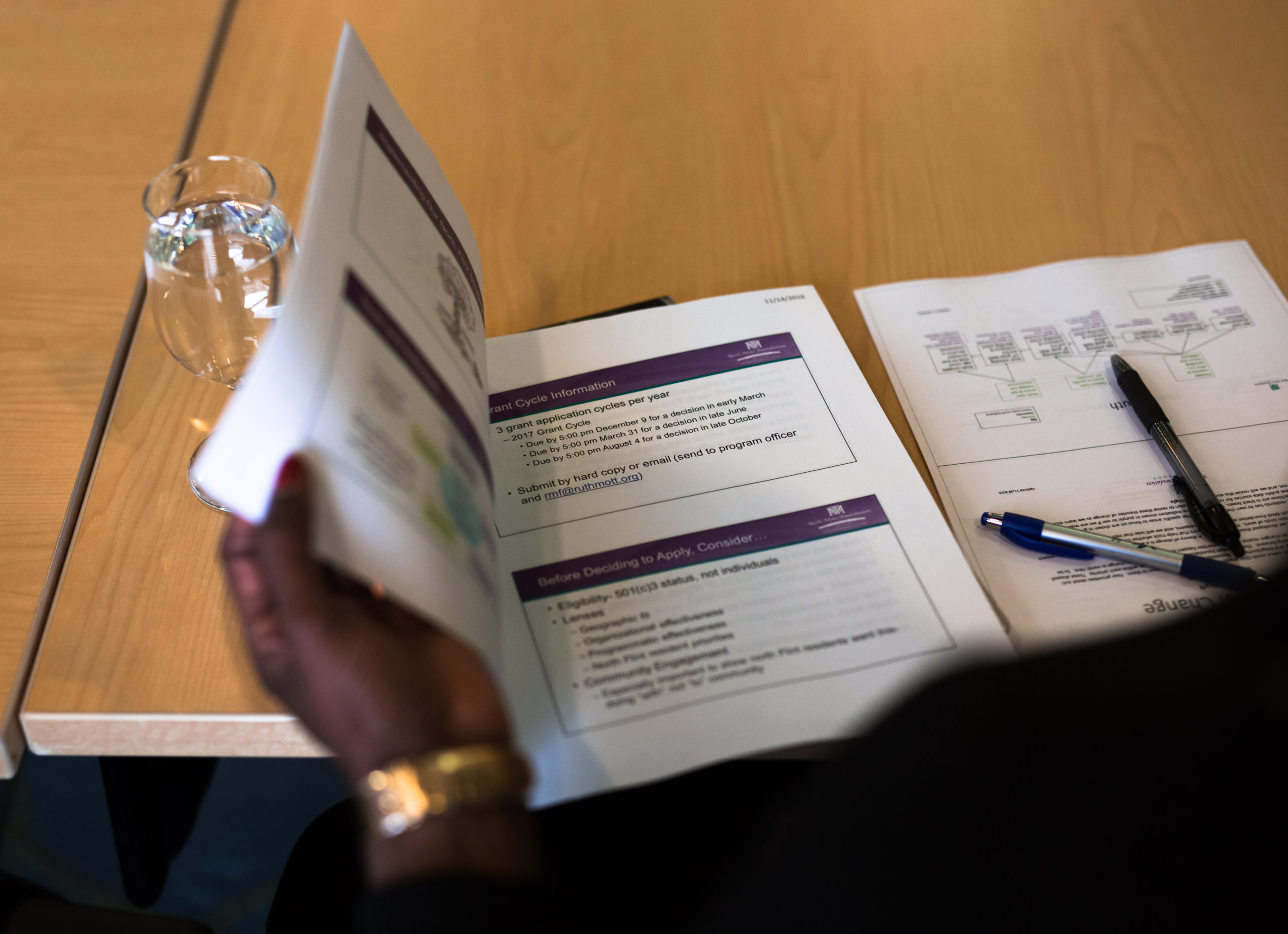 A hand is shown flipping through a stapled packet of grant guidelines on a wooden conference table.