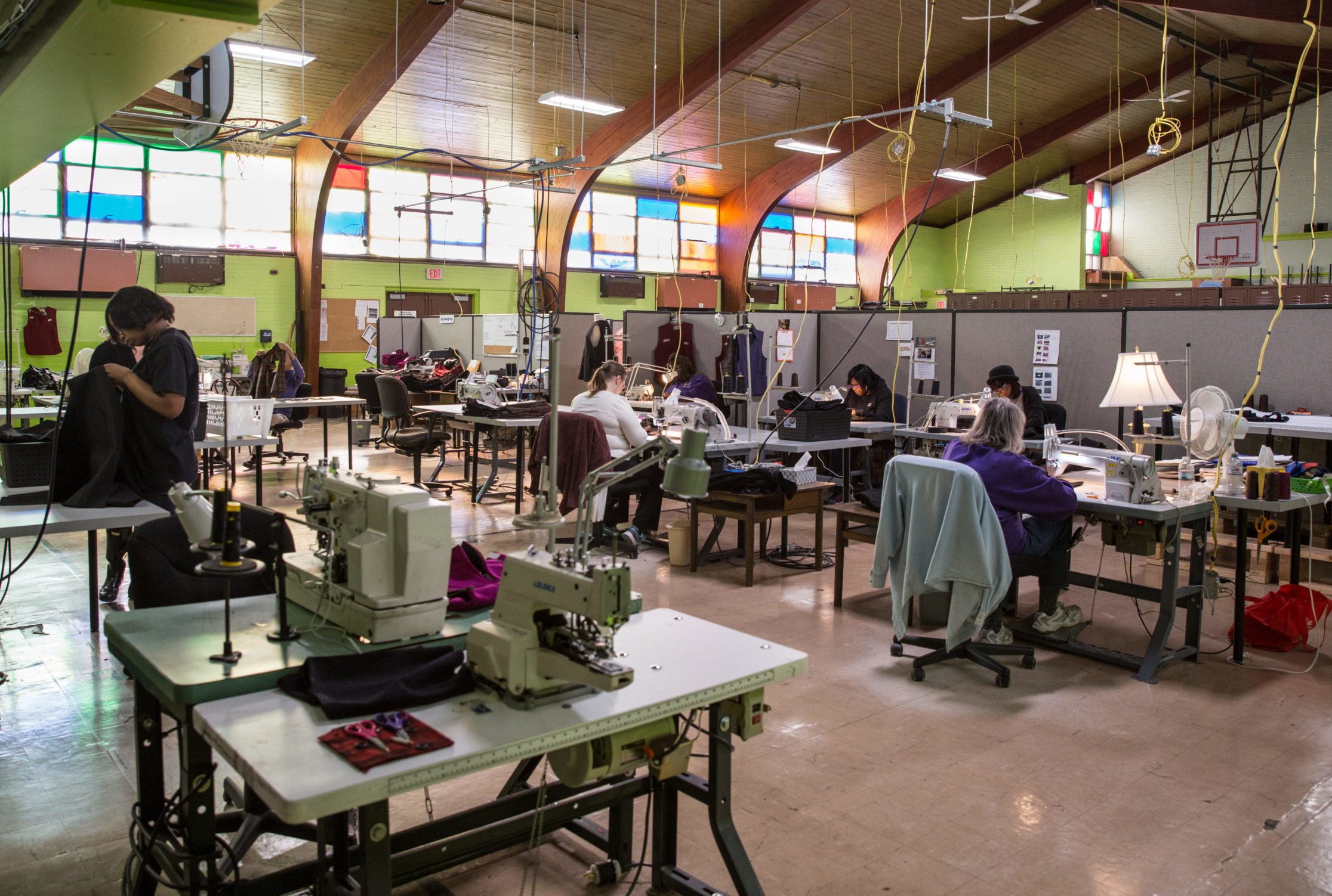 A well-lit room with stained glass windows and many people using sewing machines at St. Luke NEW Life Center