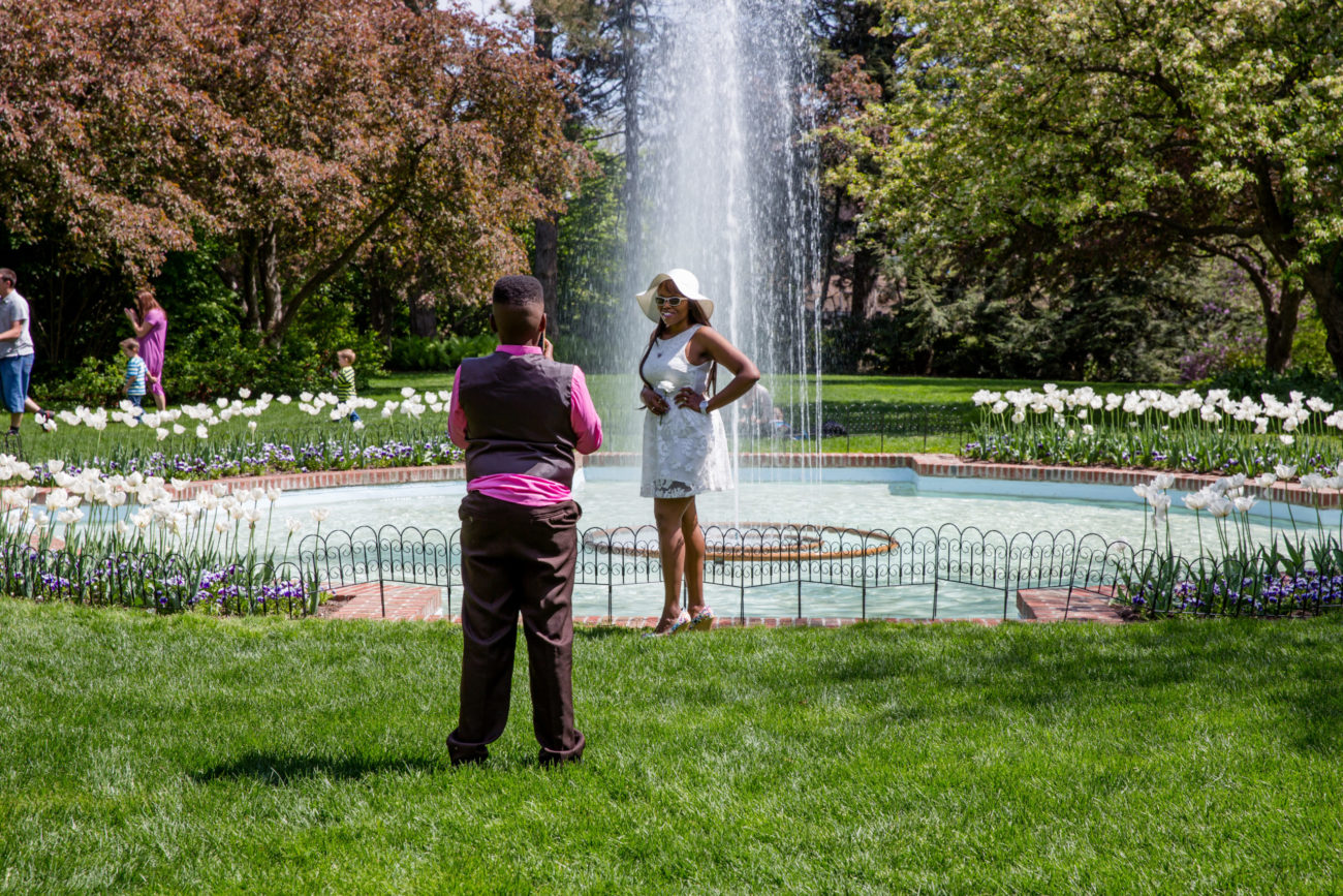 A boy takes a photo of a nicely dressed woman in front of the fountain at Applewood.