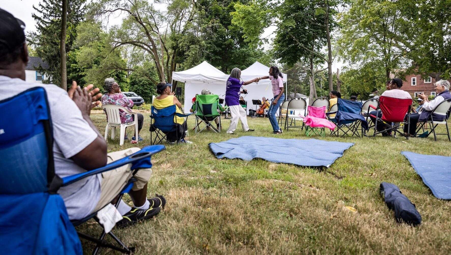 A older woman and a young woman dance on the lawn while a band plays under a tent in the background and onlookers sit in outdoor folding chairs.