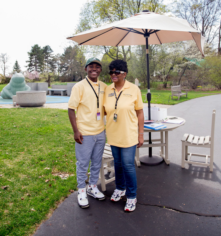 A young man wearing a yellow volunteer shirt and a woman wearing a yellow volunteer shirt pose for the camera near the entrance gate to Applewood.
