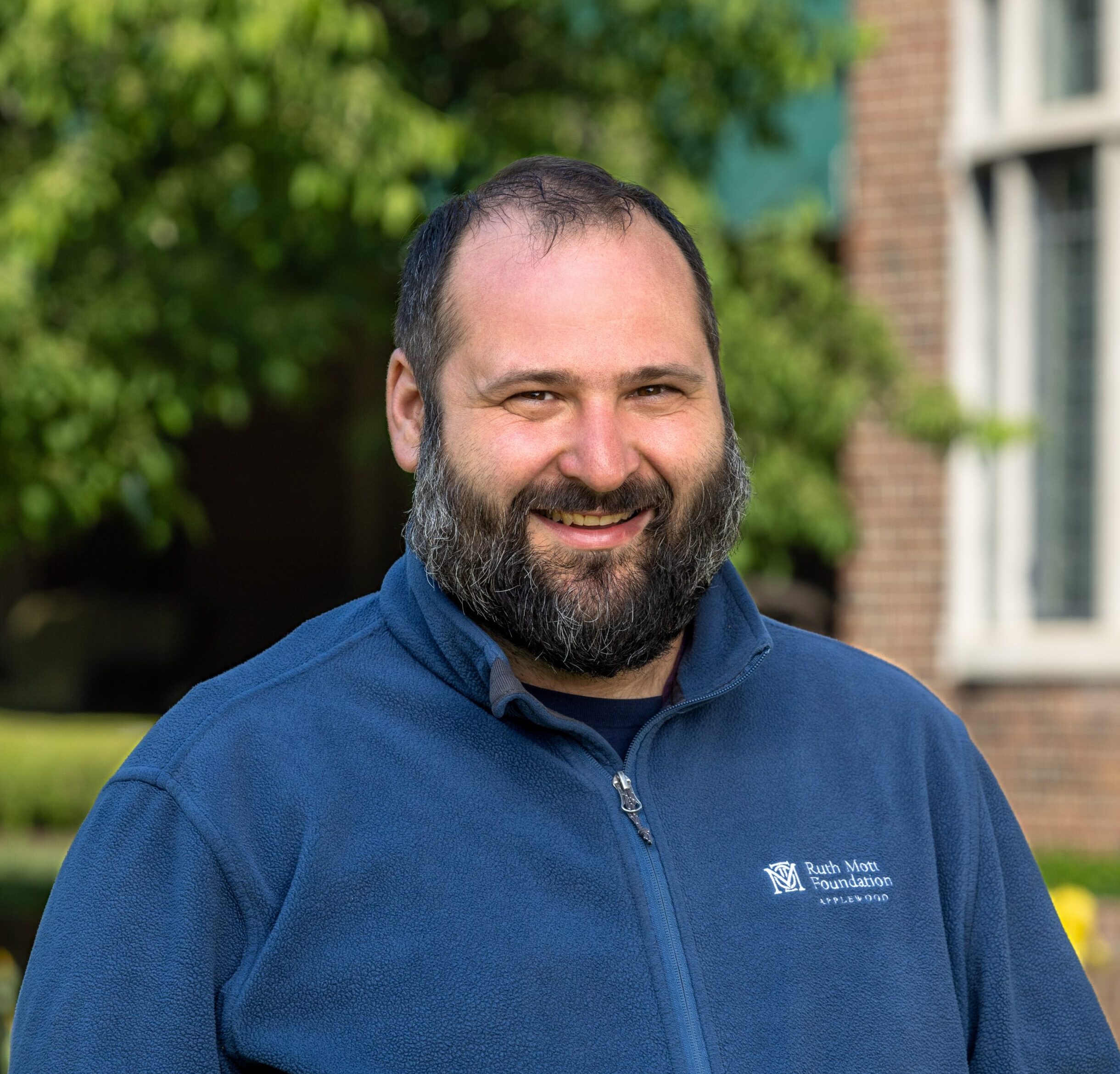 A middle-aged man with a beard, wearing a blue zip-up fleece with the Ruth Mott Foundation logo, smiles outdoors.