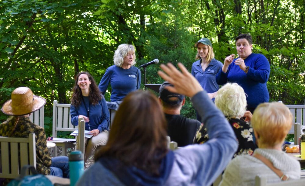 A group of people having a lunch and learn session around a table in a wooded area.