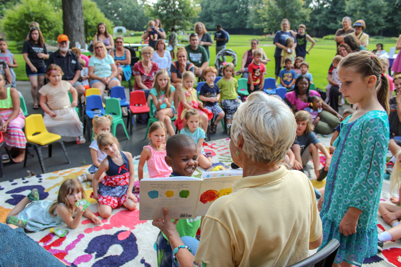 A group of young children are gathered outside on a colorful blanket. A woman not facing the camera is holding up a book during story time.