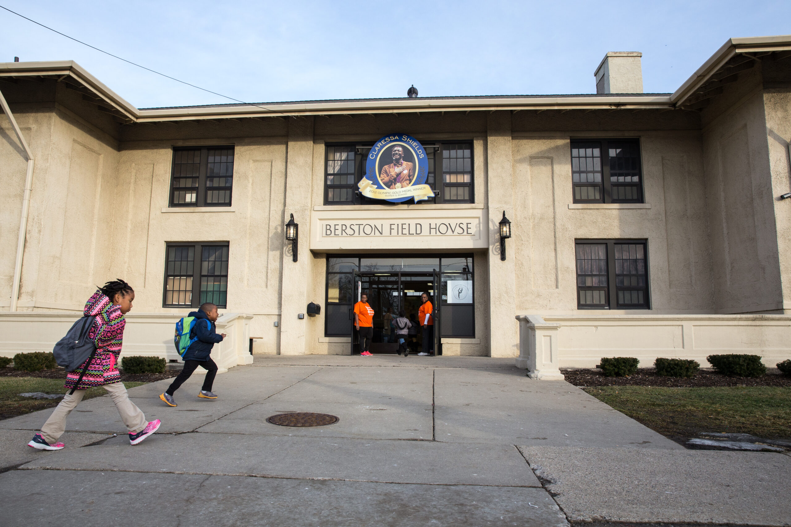 Two children with backpacks run toward the entrance of Berston Field House, pictured in the background. Adults wearing orange t-shirts hold the door open to welcome them.