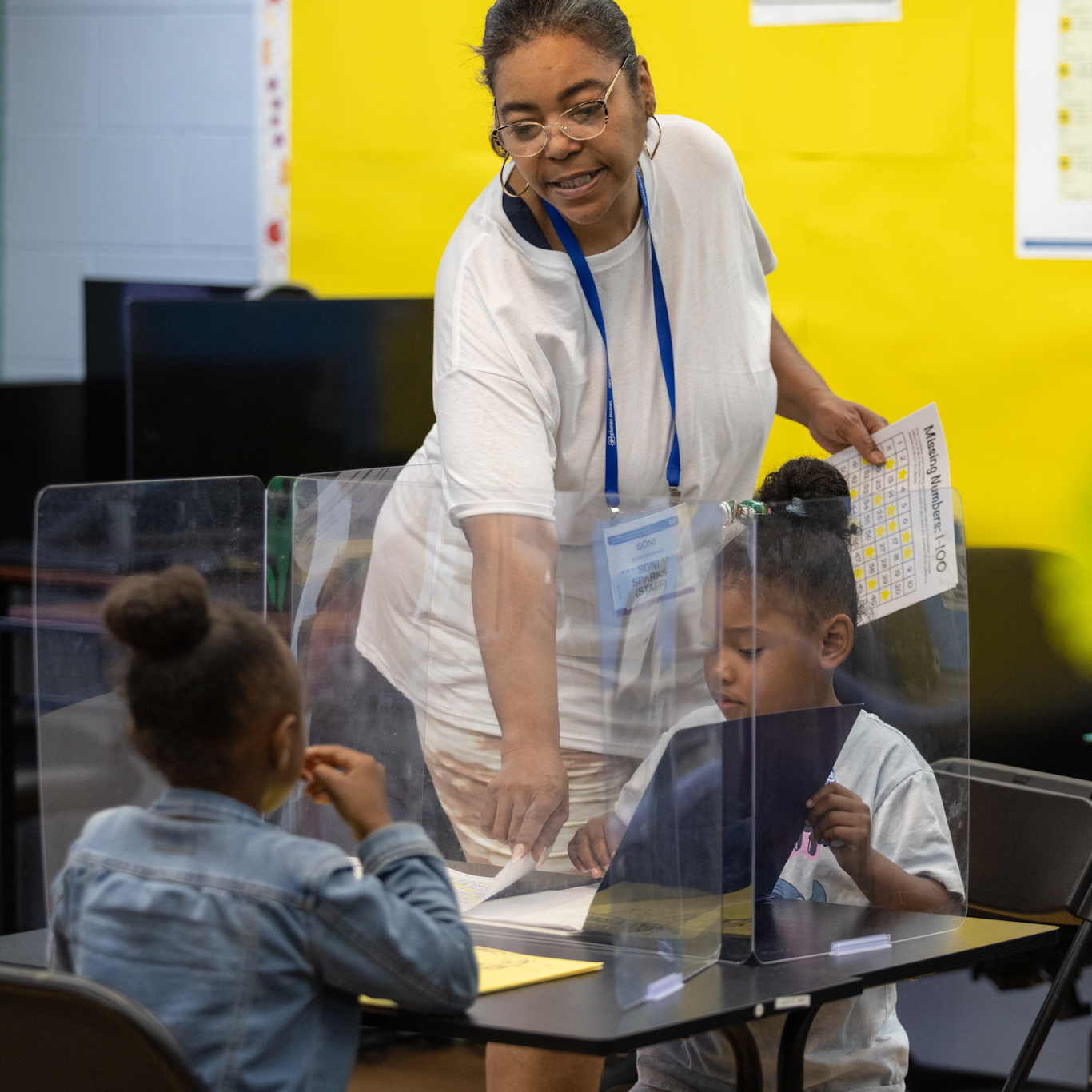 An instructor is standing and pointing to paper on a desk where two young girls are working or studying with a clear partition between them.