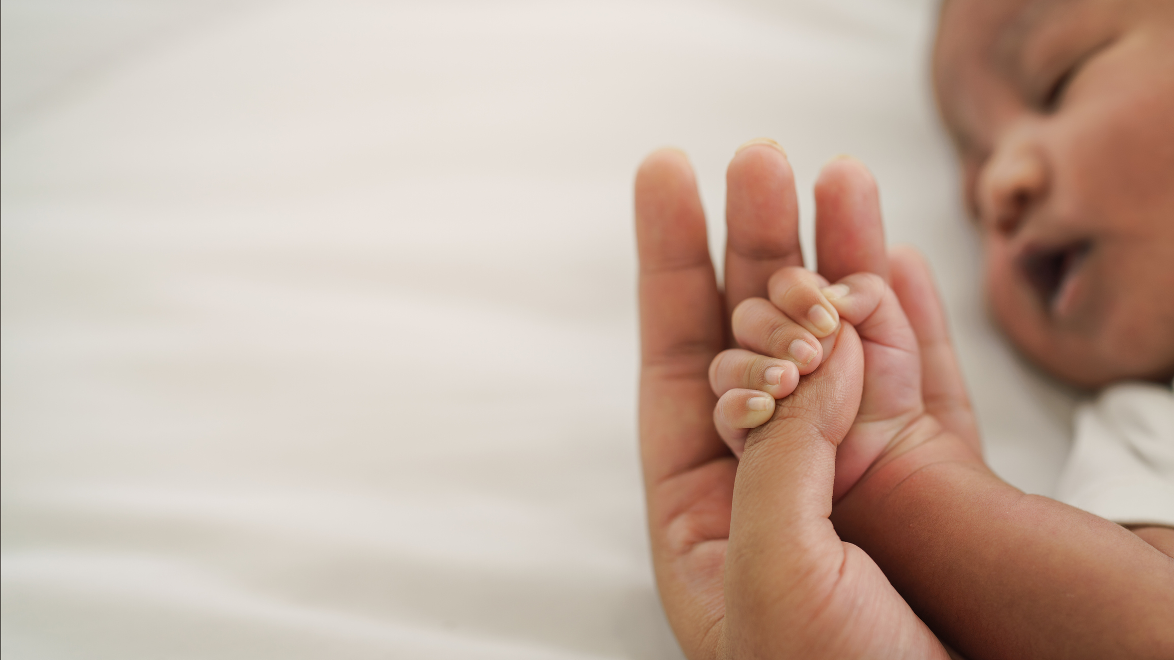 A sleeping African American baby holds the thumb of an adult whose face is off camera