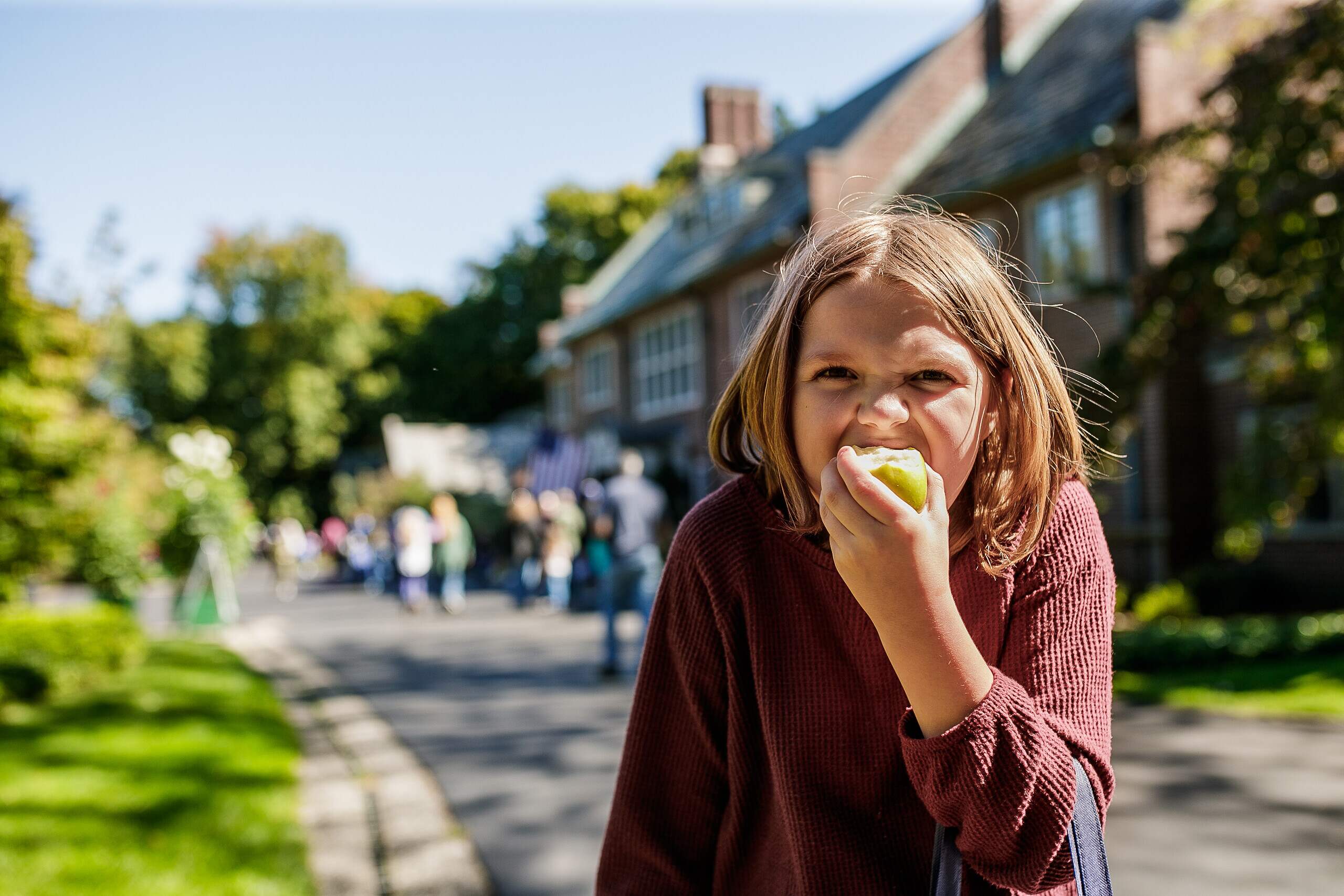 A girl bites into an Applewood. In the background, people walk by the historic home.