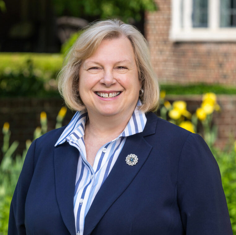 A woman with blonde hair wearing a navy blazer and striped shirt stands outside, smiling, with yellow flowers and a brick building in the background.