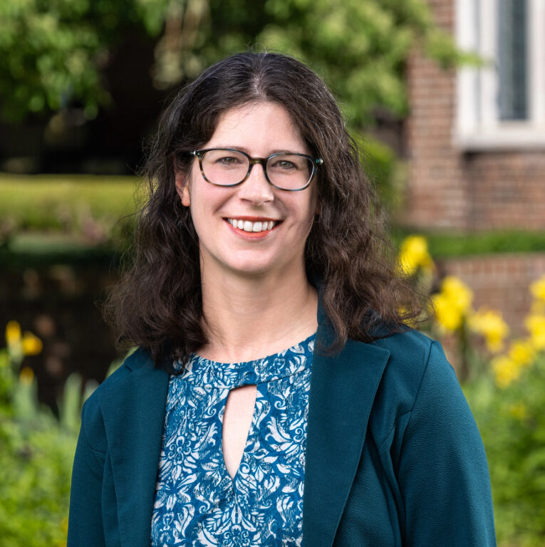 Woman with long dark hair and glasses, wearing a teal jacket and patterned blouse, smiling outdoors in a garden with yellow flowers and a brick building in the background.