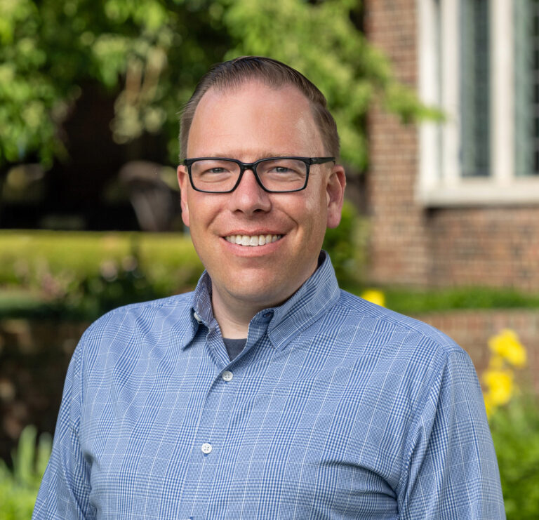 A man with glasses and a blue plaid shirt smiles while standing outside in front of a building and green plants.