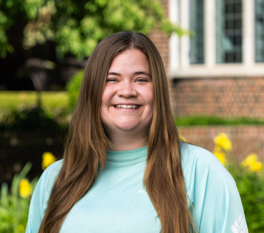 A person with long brown hair wearing a light blue shirt stands outside in front of a brick building and yellow flowers, smiling at the camera.