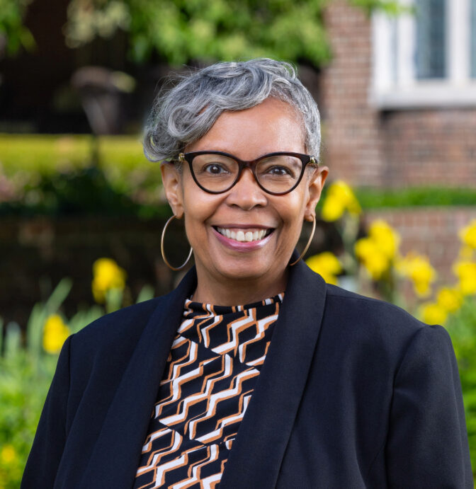 Smiling woman with short gray hair and glasses stands outdoors in front of yellow flowers and a brick building, wearing a patterned top and black blazer.