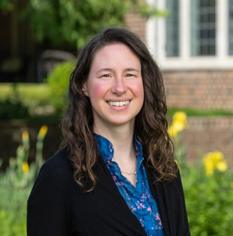 A woman with long brown hair, wearing a blue floral shirt and black cardigan, stands outside in front of greenery and a brick building, smiling at the camera.