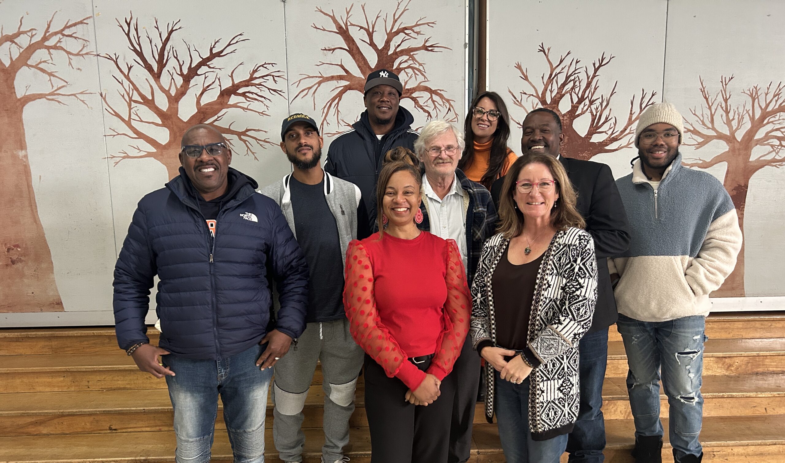 Group of eight smiling people posing for a photo in front of a wall with tree illustrations.