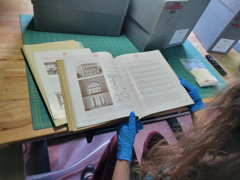 A person wearing blue gloves examines an open book on a table filled with archival materials and folders.