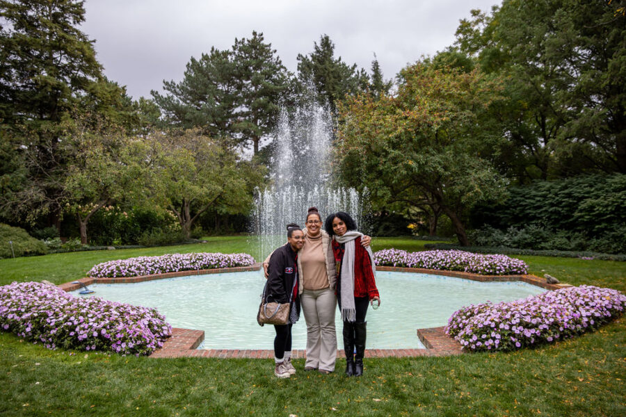 Three individuals posing for a photo in front of a fountain surrounded by flowers in a park setting.