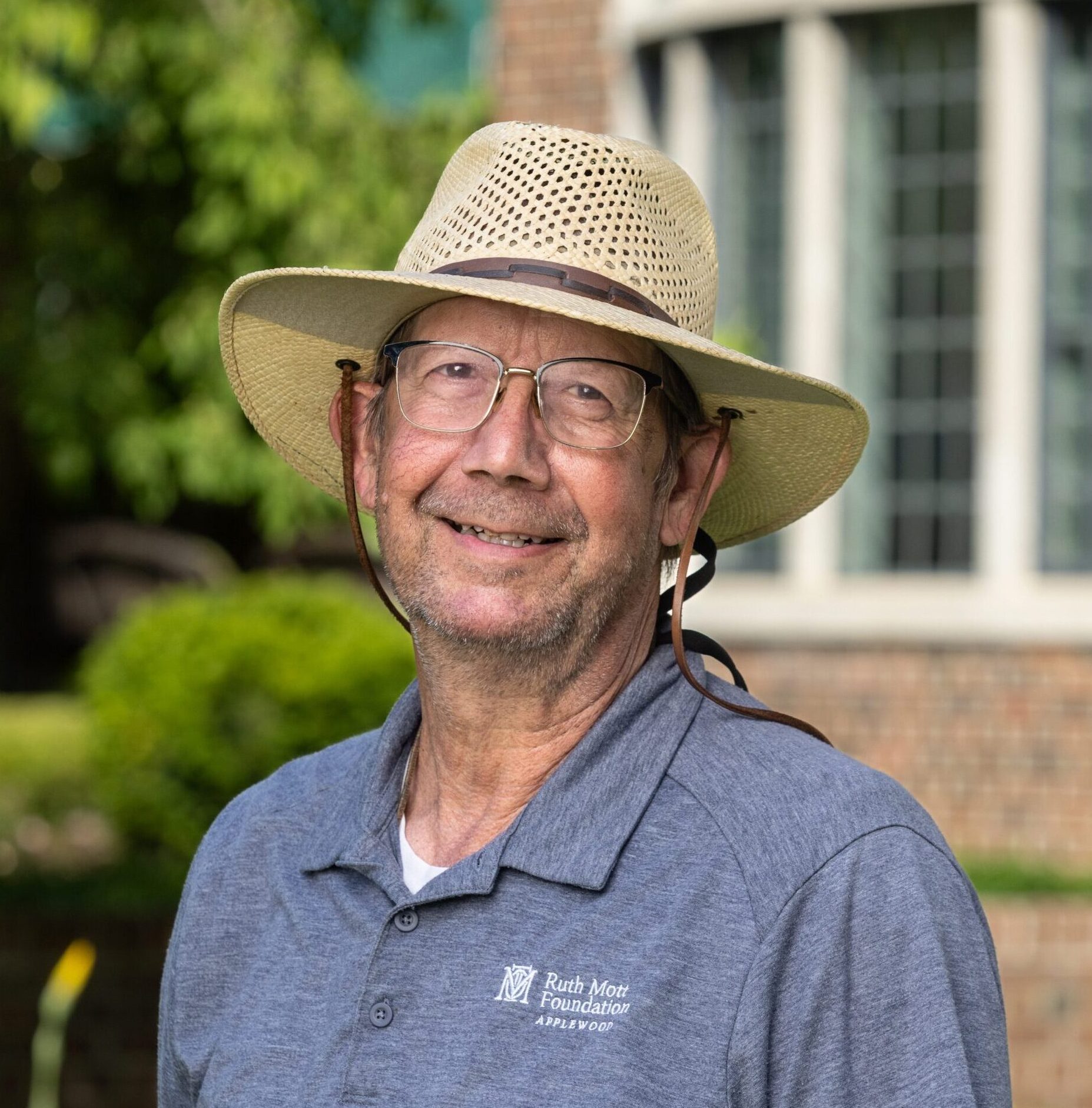 Older man wearing glasses and a straw hat, smiling outdoors in front of a brick building, dressed in a blue polo shirt with a logo.