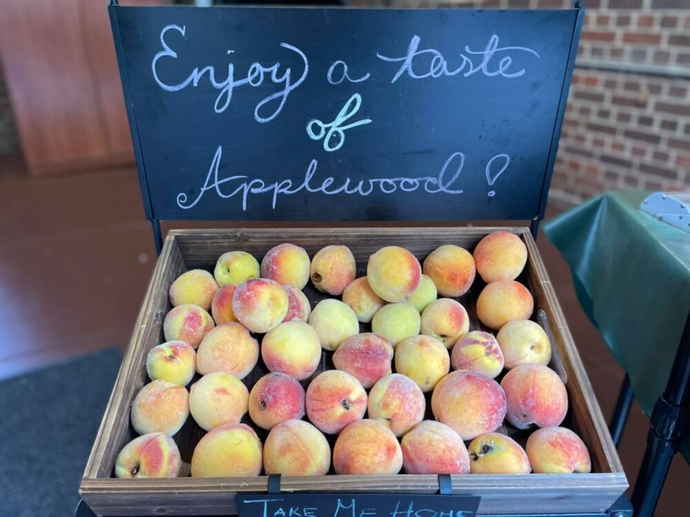 A wooden crate filled with fresh peaches sits below a sign reading “Enjoy a taste of Applewood!” at the Applewood Fall Harvest Festival.