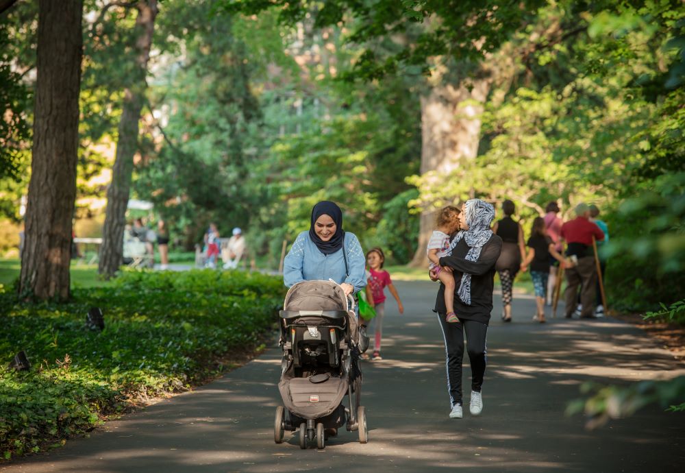 Two women, one pushing a stroller and one carrying a child, walk along a tree-lined path in a park with other people in the background—discover moments like this when you visit Applewood.