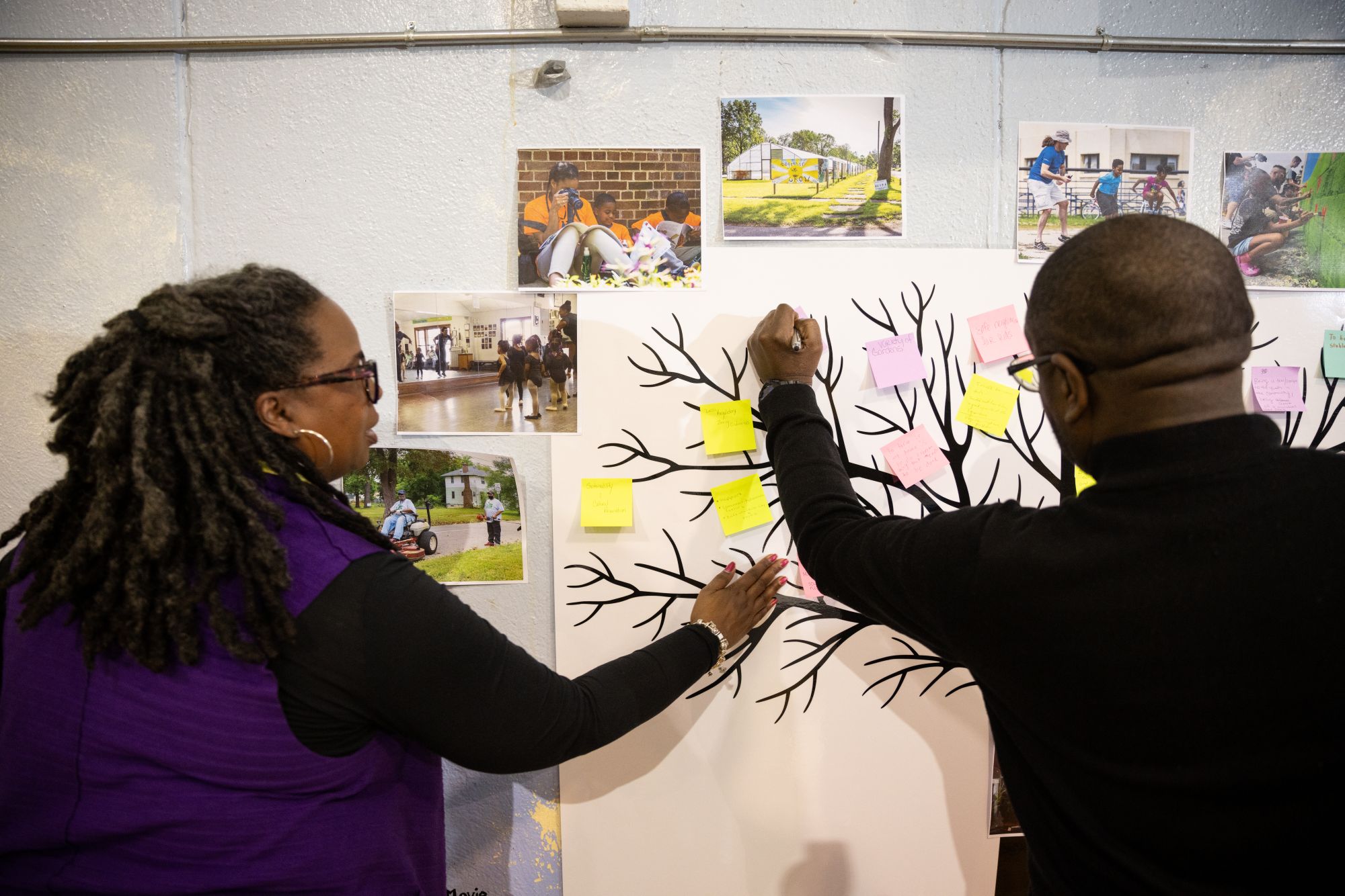 Two people place sticky notes on a tree diagram poster for the Ruth Mott Foundation, with several photographs pinned to the wall above them.