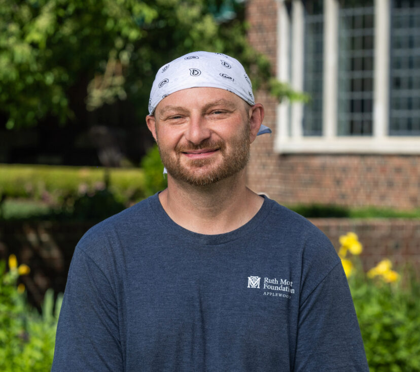 A man in a blue T-shirt and white bandana stands outdoors in front of a brick building with greenery and flowers in the background.