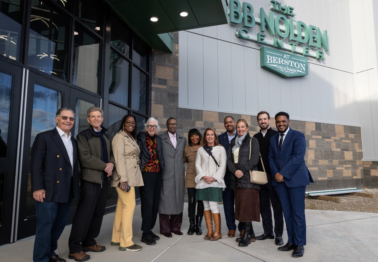 A group of eleven people, including members of the Ruth Mott Foundation Board, stand together smiling outside the entrance of the Bobby Nolden Center at Berston Field House.