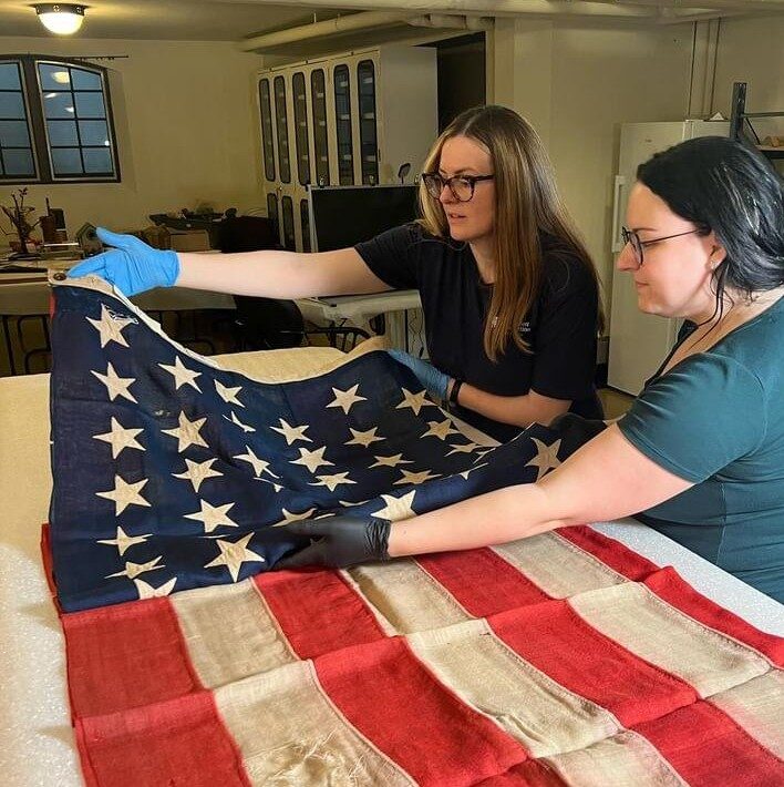 Two people wearing gloves carefully handle an old, worn American flag on a table in a well-lit room, highlighting the Ruth Mott Foundation Partnership Opportunities for preserving local history.