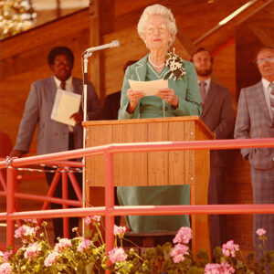 An elderly Ruth Mott stands at a podium reading from papers, with three men in suits behind her and pink flowers in front of the stage.