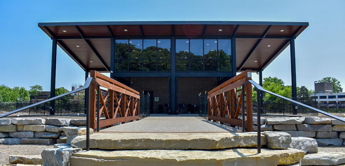A modern pavilion with a flat roof, part of the Applewood Estate Master Plan, is seen from the front, approached by a stone stairway and a metal-railed bridge, with trees and buildings in the background.