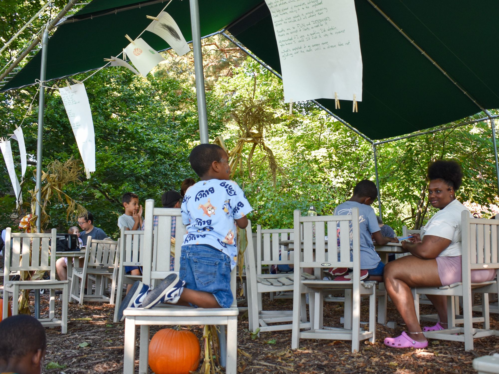 Several children sit and work at tables under a tent outdoors, with papers hanging overhead and a pumpkin on the ground in front. One child kneels on a chair, looking up—an inviting scene to Plan Your Visit to Applewood.