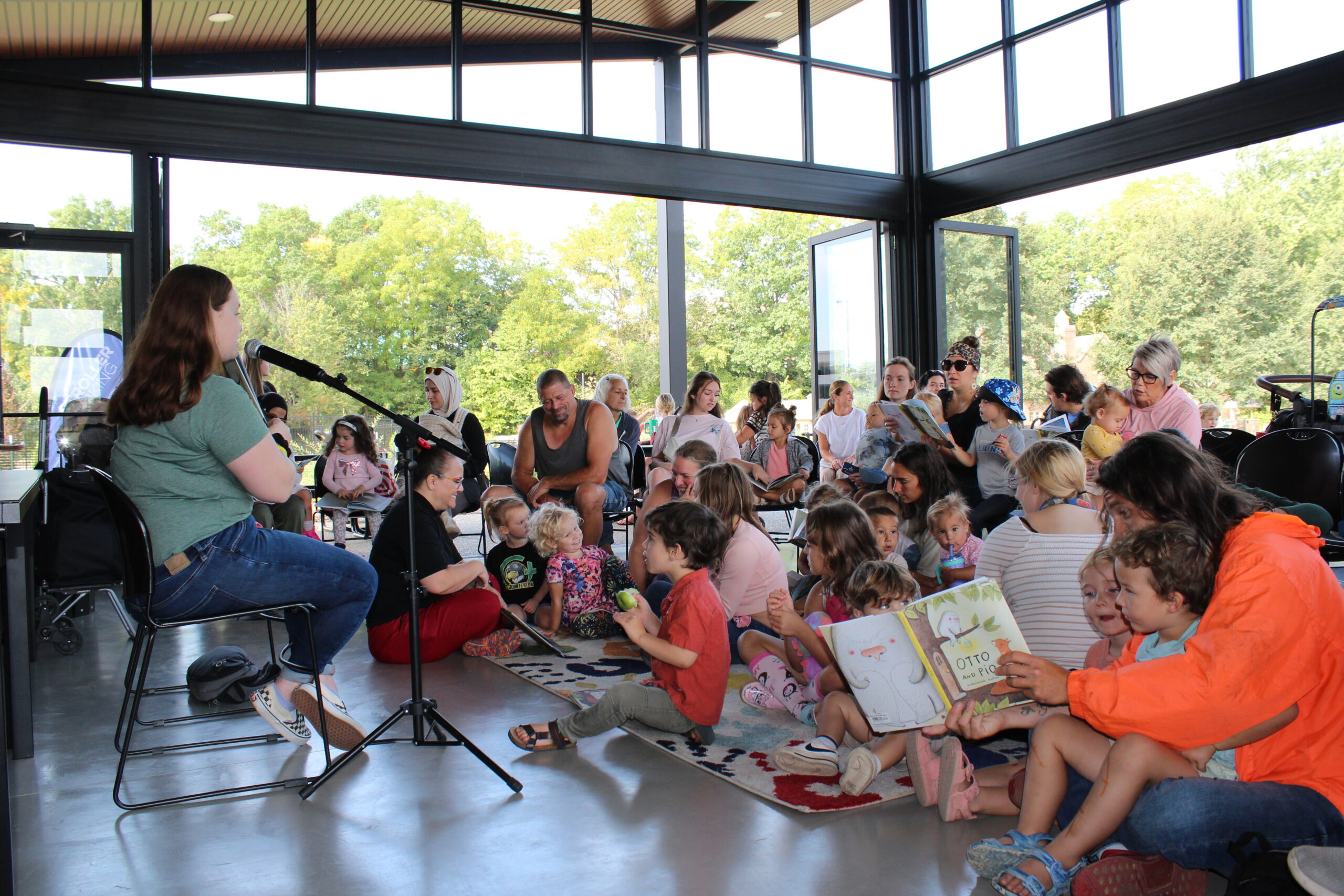 A woman sits at a microphone reading to a group of children and adults gathered on the floor and in chairs in a bright room with large windows.