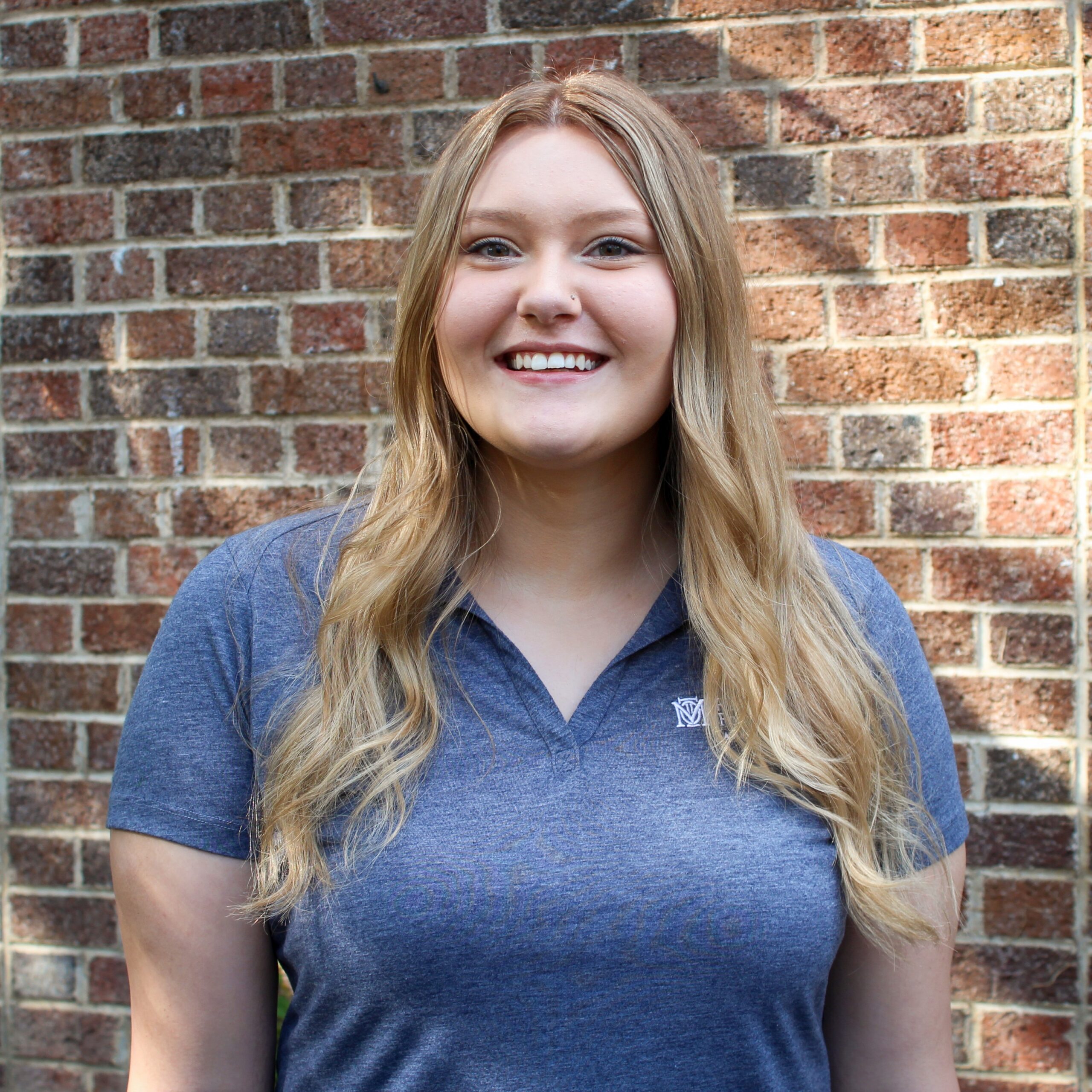 A woman with long blonde hair wearing a blue collared shirt stands in front of a brick wall, smiling at the camera.