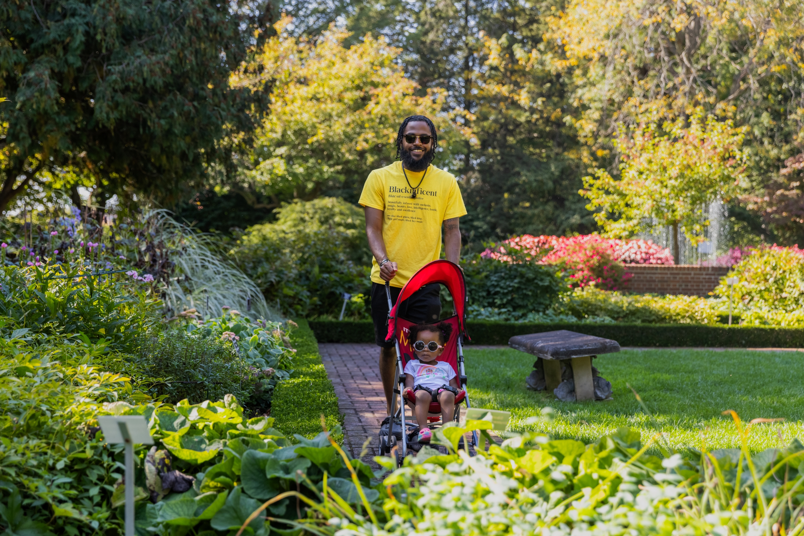 An adult pushes a child in a stroller along a garden path at the Applewood Estate, surrounded by greenery, flowers, and trees on a sunny day—a scene nurtured by the Ruth Mott Foundation.