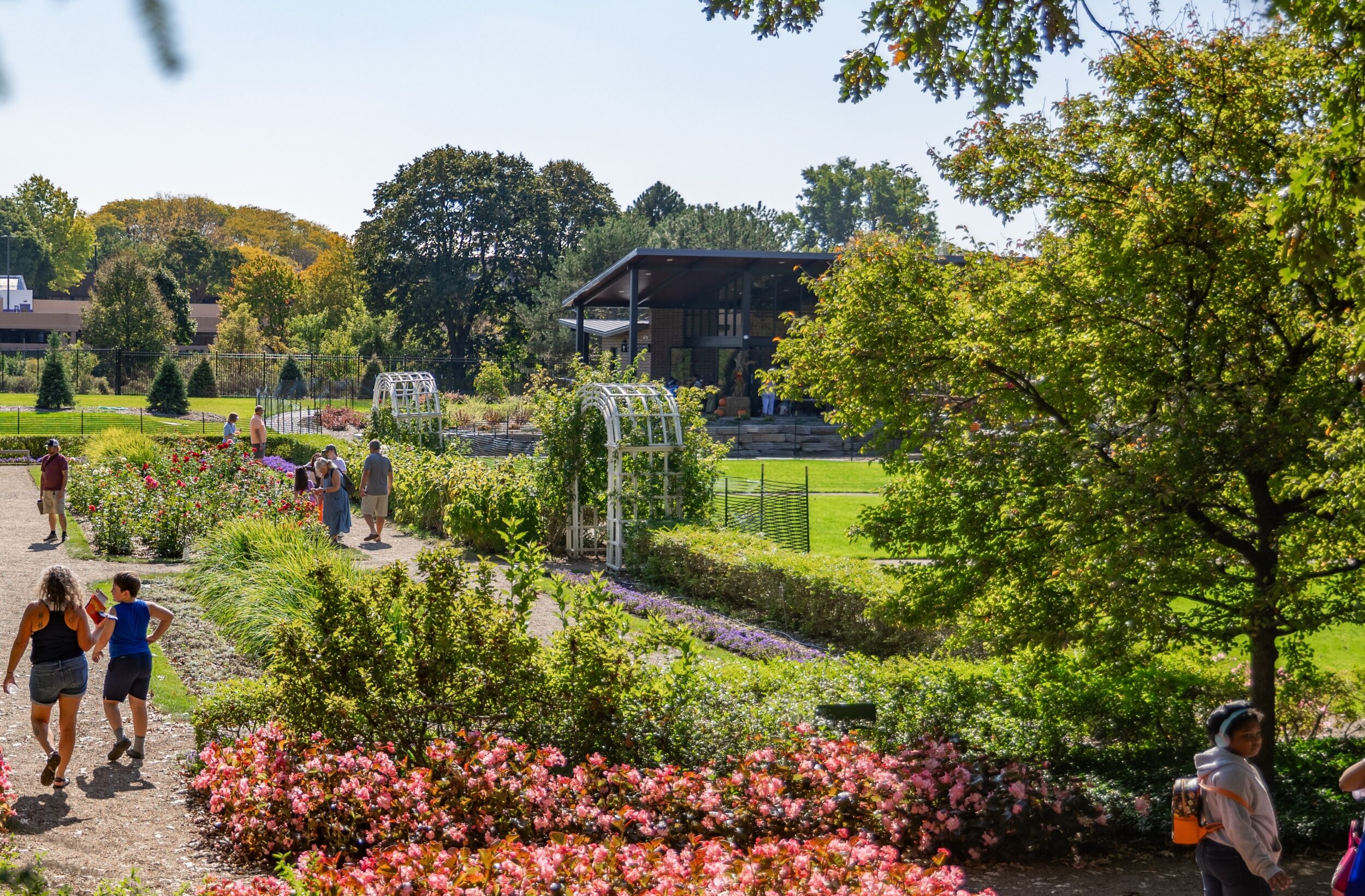 People walk along garden paths surrounded by blooming flowers, green trees, and a pavilion in the background on a sunny day.