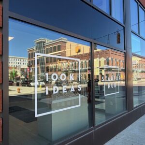 Downtown Flint buildings and street are reflected in a glass window with the 100K Ideas logo on it.
