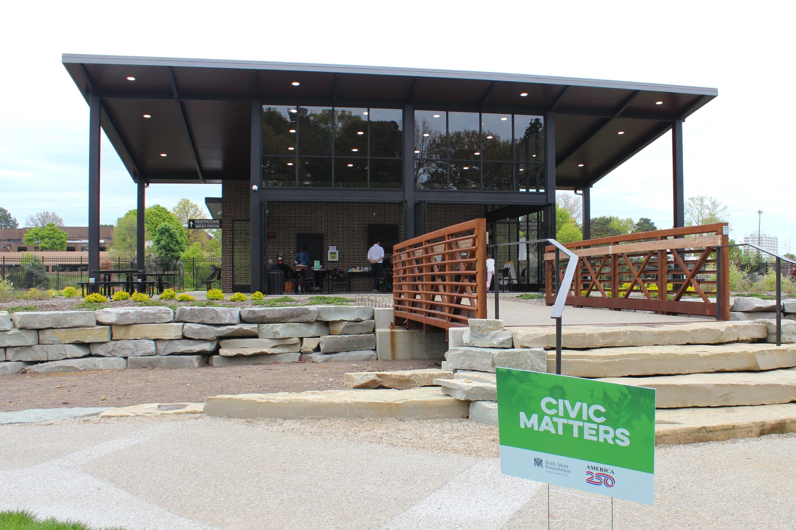 A modern pavilion with glass walls and a wooden bridge, featuring a sign in the foreground that reads 