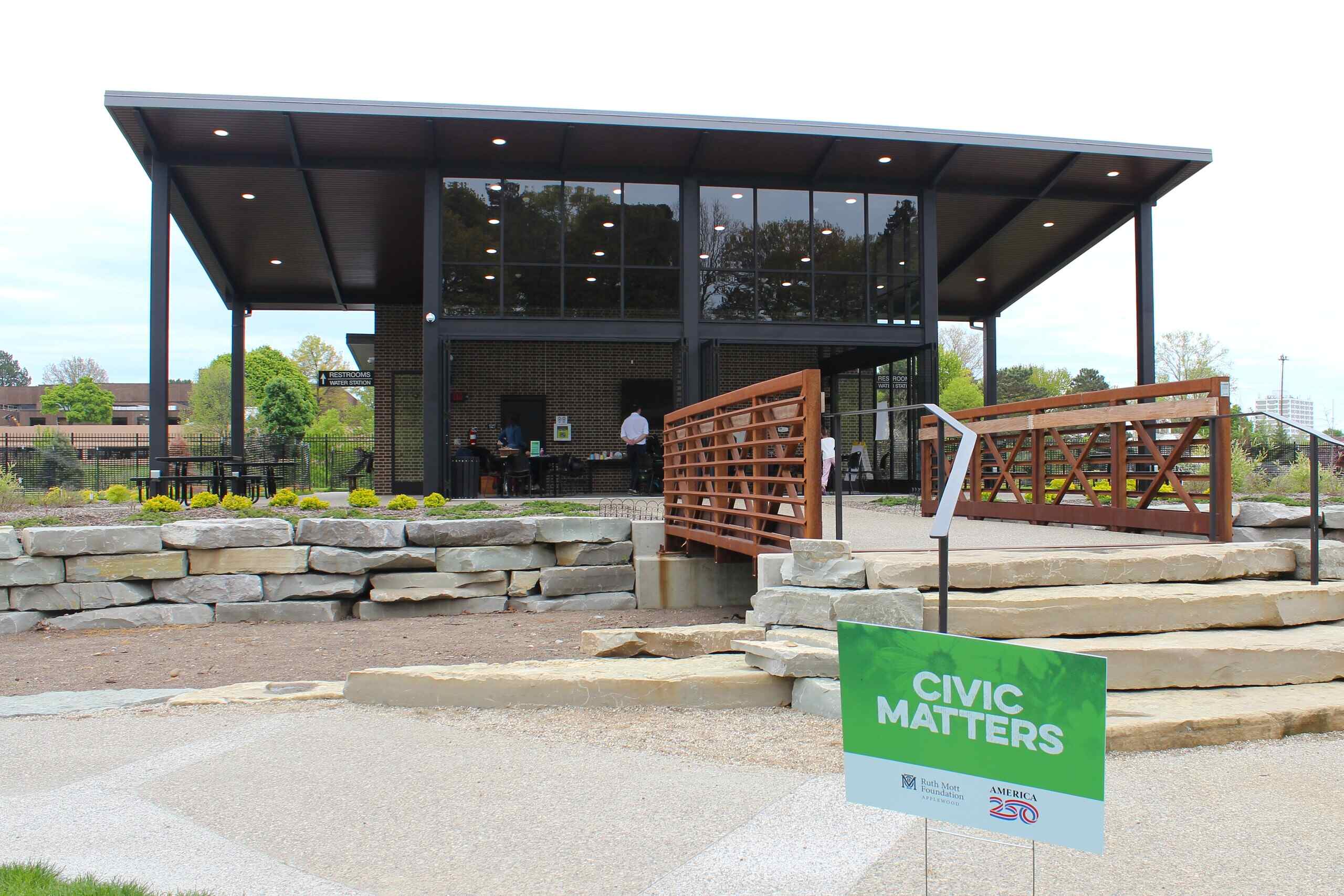 A modern pavilion with glass walls and a wooden bridge, featuring a sign in the foreground that reads 