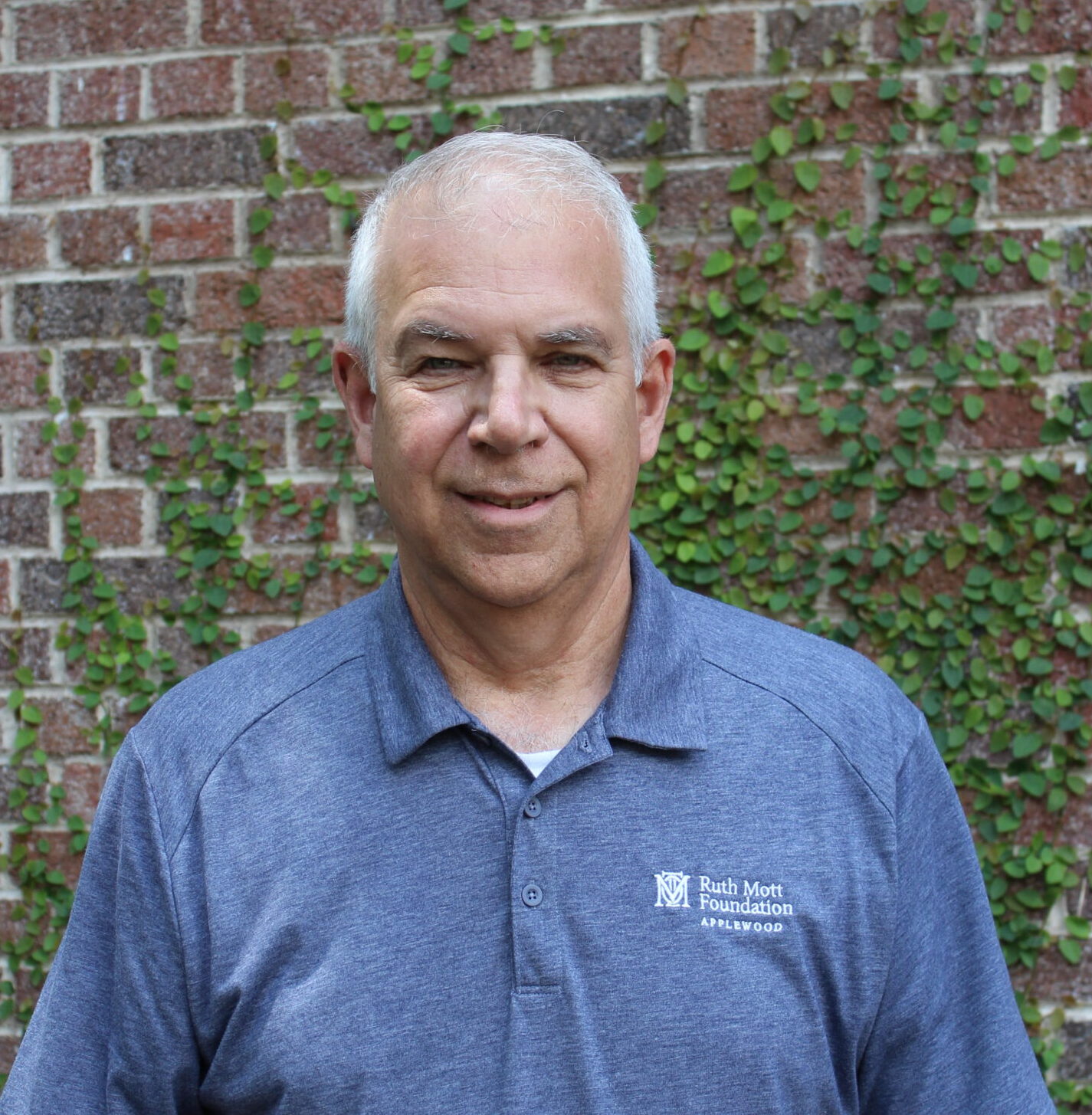 A man with short gray hair stands in front of a brick wall with green vines, wearing a blue polo shirt with a small logo on the chest.
