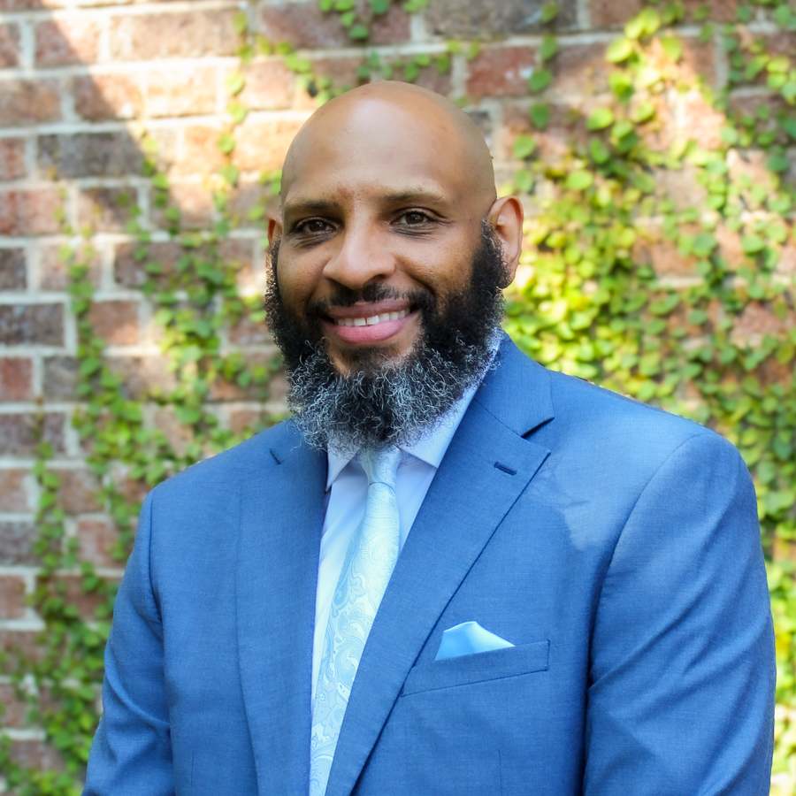 A man with a bald head and a beard, wearing a light blue suit and tie, stands smiling in front of a brick wall with green ivy.
