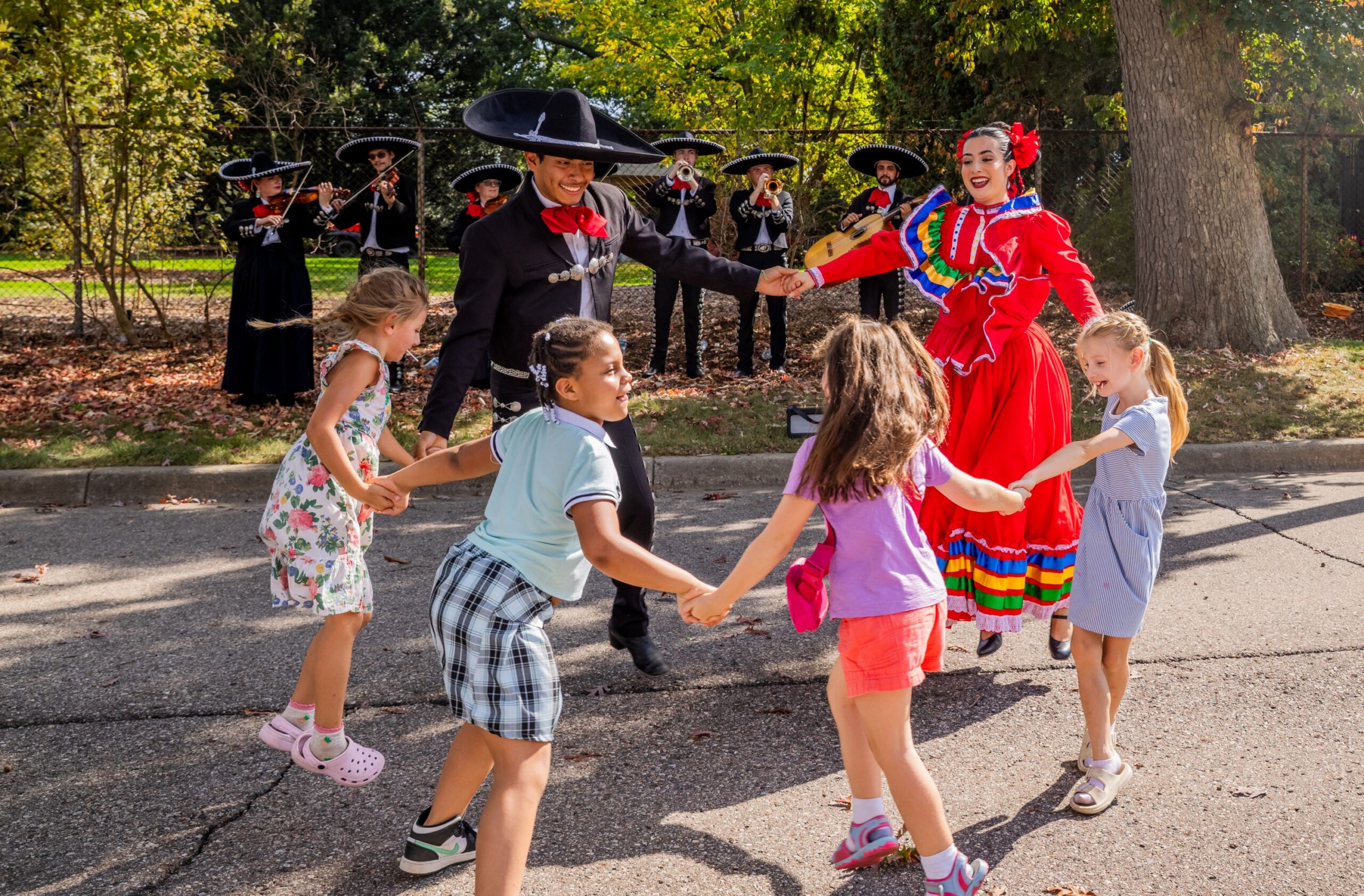 Four children hold hands and dance in a circle with two adults in traditional Mexican attire, while a mariachi band plays in the background outdoors at a Ruth Mott Foundation event.